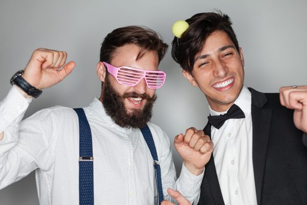 Cheerful young multiethnic male best friends in elegant clothes and party glasses and headband smiling happily and clenching fists in dance against gray background